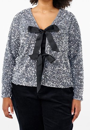 Haut à manches longues en sequins argentés avec deux grands nœuds en satin noir, porté sur une chemise noire et associé à un pantalon noir.