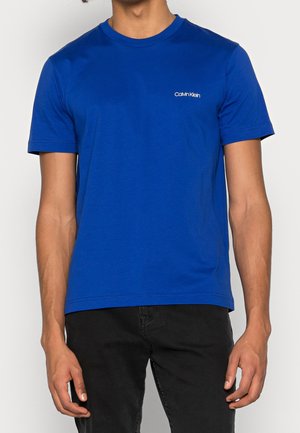 Homme portant un t-shirt bleu uni Calvin Klein et un pantalon noir, debout devant un fond neutre.