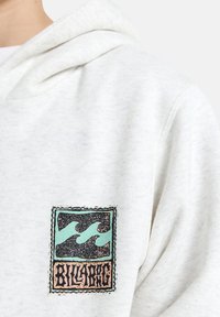 Lichtgrijze hoodie met een vierkant geborduurd patch met turquoise golven en zwarte accenten, omrand met een decoratieve rand.