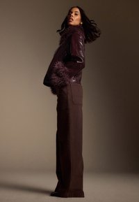 Giacca in pelle bordeaux con colletto e polsini morbidi, abbinata a pantaloni marroni a gamba larga. Texture liscia e design aderente con accenti distinti.