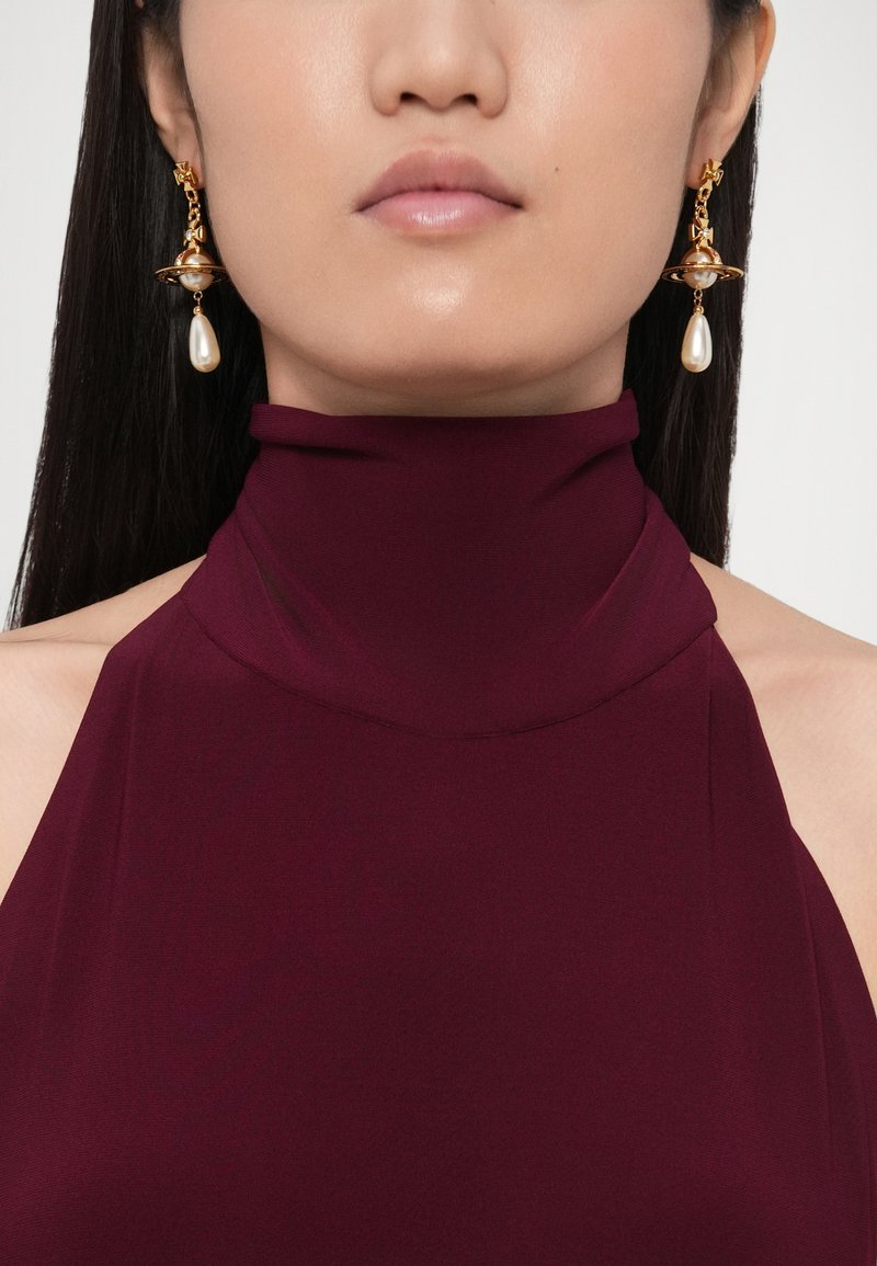 Haut col roulé bordeaux à texture lisse, associé à des boucles d'oreilles dorées ornées d'une perle et d'un motif orbital complexe.