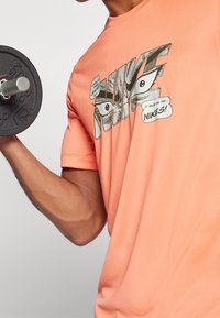 Koralfarvet Nike T-shirt med korte ærmer, prydet med et grafisk design med øjne og tekst. Modellen holder en sort vægt. Glat stoftekstur.