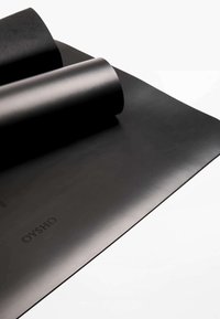 Tapis de yoga noir partiellement ouvert, fabriqué en matériau lisse avec un subtil éclat ; marqué du logo "OYSHO" dans le coin inférieur gauche.