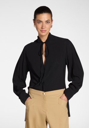 Vrouw draagt een zwart blouse met lange mouwen en een strik bij de hals, gecombineerd met beige, hooggetailleerde broek en heeft haar handen in de zakken.