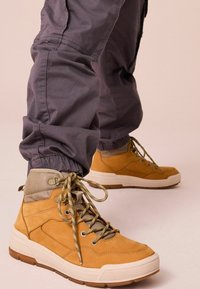 Brązowe skórzane sneakersy typu high-top z szarymi tekstylnymi akcentami, płaskimi sznurowadłami i masywnymi białymi gumowymi podeszwami. Noszone z szarymi zwężającymi się spodniami.