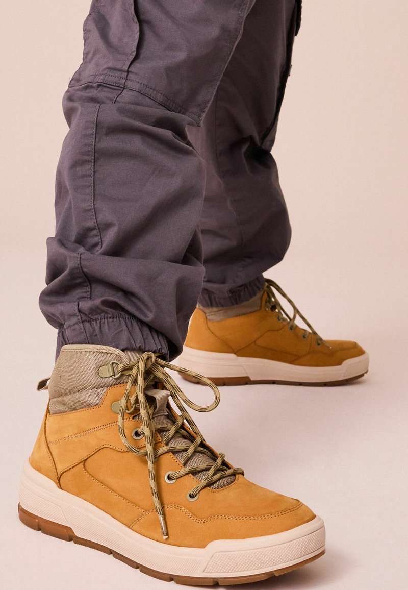 Brązowe skórzane sneakersy typu high-top z szarymi tekstylnymi akcentami, płaskimi sznurowadłami i masywnymi białymi gumowymi podeszwami. Noszone z szarymi zwężającymi się spodniami.