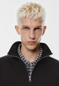 Svart pullover med dragkedja och ribbad krage över en rutmönstrad skjorta med knappar. Ljusfärgat hår, neutral bakgrund.