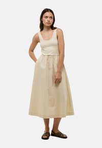Robe maxi beige clair. Corsage ajusté avec de larges bretelles, taille froncée et jupe fluide. Portée avec des sandales en cuir marron à lanières.
