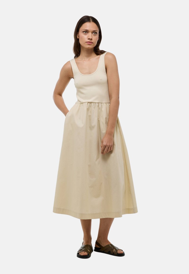 Robe maxi beige clair. Corsage ajusté avec de larges bretelles, taille froncée et jupe fluide. Portée avec des sandales en cuir marron à lanières.