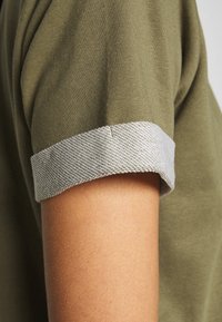 Olivewą zielona koszulka z rękawem, z złożonym jasnoszarym teksturowanym mankietem, prezentująca gładki materiał i dopasowany krój.