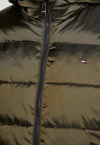 Tmavě zelená prošívaná bunda s lesklou texturou, s černým zipem a malou nášivkou s logem v červené, bílé a modré barvě na hrudi.