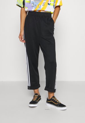 Person iført sorte Adidas træningsbukser med hvide striber og sorte sneakers med farverige snørebånd, stående med den ene hånd i lommen.