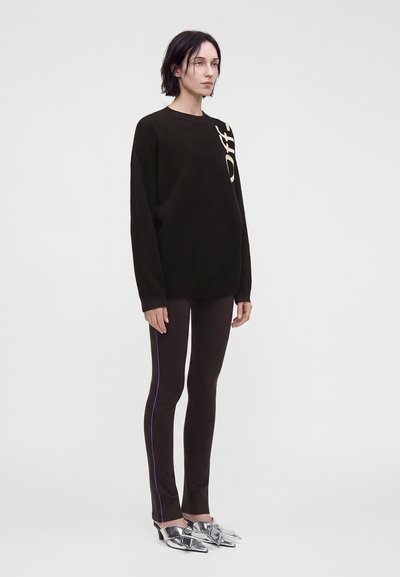 Femme aux cheveux courts et foncés portant un pull noir oversize avec un texte blanc partiel, un pantalon noir avec des bandes latérales violettes et des talons pointus argentés.