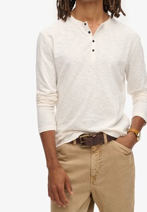 Homme portant une chemise henley à manches longues crème avec des boutons noirs, un pantalon beige, une ceinture en cuir marron et une montre-bracelet en or, la main dans la poche.
