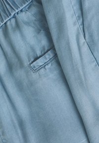 Světle modrý denimový materiál s hladkou strukturou, nařaseným pasem a malou náprsní kapsou s kontrastním švem.