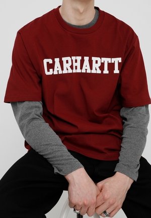 Maroon short-sleeve t-shirt featuring bold white "CARHARTT" text, layered over a gray long-sleeve shirt. Cotton fabric, casual fit.