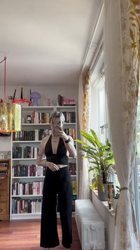 Svart ribbad halternecktopp kombinerad med vida svarta byxor. Kort ljust hår och synliga tatueringar. Speglar och växter i bakgrunden.