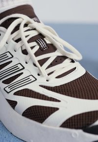 Sneaker marron avec des accents blancs, tige en mesh, lacets ronds et semelle en caoutchouc texturée. Présente des lignes de design sur les côtés.