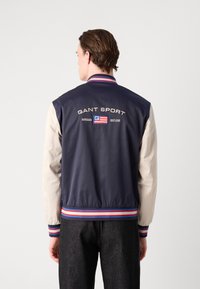 Marinblå och krämfärgad bomberjacka med ribbat krage och muddar, med "GANT SPORT"-logotyp och flaggdetalj på baksidan.