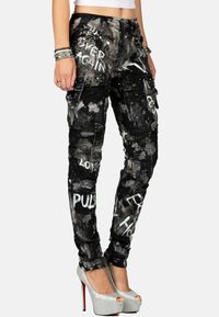 Zwarte cargobroek met abstracte witte verfsproeien en tekst. Bevat meerdere zakken en decoratieve studs. Gecombineerd met zilveren hoge hakken.