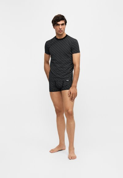 Homme debout pieds nus portant un haut noir à manches courtes assorti et un short avec un motif de texte diagonal blanc sur un fond clair uni.