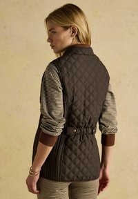 Kvadratuveida brūnā veste ar jostas loksni, kurai ir kontrastējošas houndstooth raksta piedurknes un ribotās brūnās aproces.