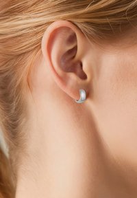 Orecchino a cerchio in argento con una finitura liscia e lucida. Il design è curvo e avvolge parzialmente il lobo dell'orecchio. La vista ravvicinata mette in evidenza la texture.