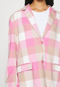 Femme portant un blazer à grands carreaux rose, beige et blanc sur un top blanc, avec deux poches à rabat à l'avant et des boutons roses.