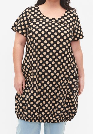 Femme portant une robe noire mi-longue à pois beige, manches courtes et ourlet élastique, associée à un jean bleu clair.