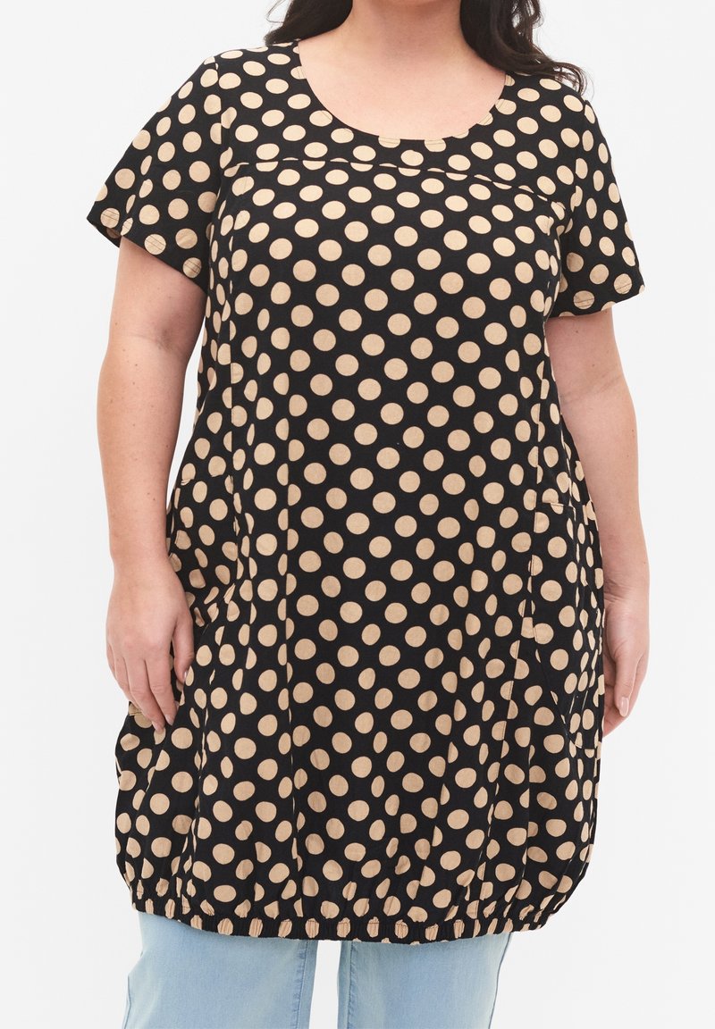 Femme portant une robe noire mi-longue à pois beige, manches courtes et ourlet élastique, associée à un jean bleu clair.