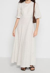 Femme portant une longue robe blanche à fines rayures avec des manches bouffantes, portant un sac à bandoulière marron et des sandales ouvertes.