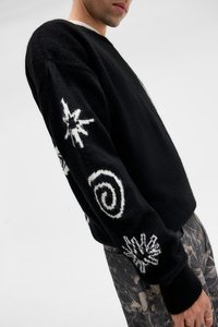 Μαύρος πλεκτός πουλόβερ με λευκά γραφικά σχέδια, συμπεριλαμβανομένων αστεριών και στροβιλισμών, με στρογγυλή λαιμόκοψη και ριχτές μανσέτες.