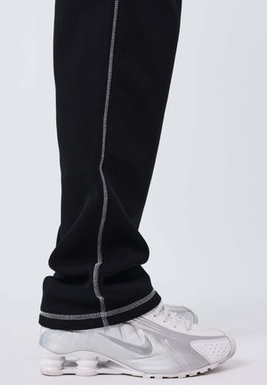 Pantaloni neri con cuciture bianche che coprono la caviglia sono indossati sopra sneaker Nike bianche e argentate con suole visibili simili a molle.