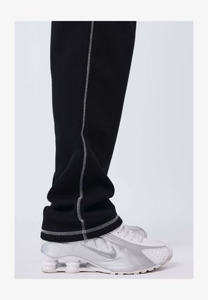 Un pantalon noir avec des coutures blanches couvre la cheville, posé sur des baskets Nike blanches et argentées avec des semelles visibles ressemblant à des ressorts.