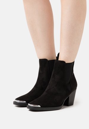 Bottines en daim noir avec un bout pointu, des panneaux latéraux élastiques et un accent métallique argenté sur le bout. Talon empilé pour plus de hauteur.