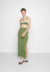 Conjunto de malha verde em duas peças, composto por um sutiã cropped com um detalhe branco, saia de cintura alta com uma fenda lateral e sandálias brancas com tiras.