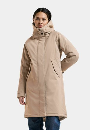 Kvinde iført en beige lang vinterjakke med høj krave, frontlynlås og klaplommer, står med den ene hånd bag på ryggen.