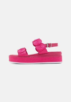 Pink plateau-sandal med flettede teksturerede remme, cirkulære dekorative elementer, ankelrem og spædelukning på hvid baggrund.