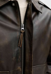 Közeli kép egy sötétbarna bőrdzsekiről, melynek ezüstcipzárja részben nyitva van, alatt világos bézs színű kötött pulóver látható.