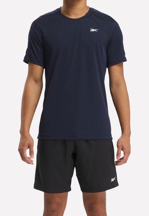Mann trägt ein marineblaues kurzärmeliges Sportshirt und schwarze Shorts mit kleinen weißen Markenlogos, steht vor einem einfarbigen Hintergrund.