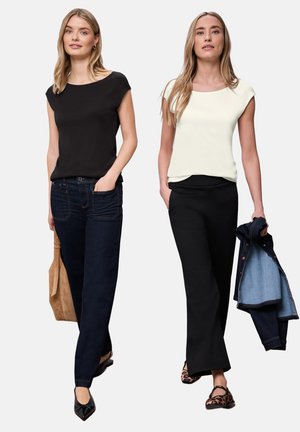 To kvinder går, den ene iført en sort top og mørke jeans med en tanfarvet taske, den anden i en hvid top og sorte bukser, der holder en denimjakke.