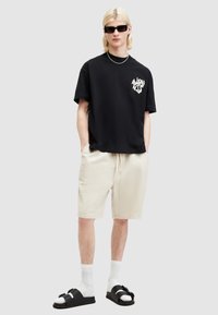 Tricou negru din bumbac cu un grafic alb, pantaloni scurți bej dintr-un material ușor, sandale negre cu detaliu sub formă de fundă și șosete albe.