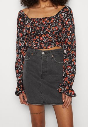 Vrouw die een zwarte bloemenprint off-shoulder top met lange mouwen en een hooggetailleerde donkergrijze spijkerbroek minirok draagt, staand voor een effen achtergrond.