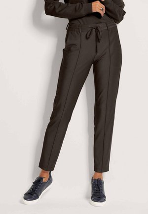 Vrouw draagt nauwsluitende zwarte taps toelopende broek met voorste naaddetails en zwarte sneakers met witte zolen, staand tegen een effen achtergrond.