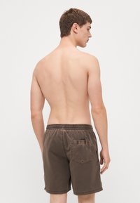 Brune svømmebukser med elastisk talje, baglomme og syet kant. Stofferne har en glat tekstur og et afslappet design.