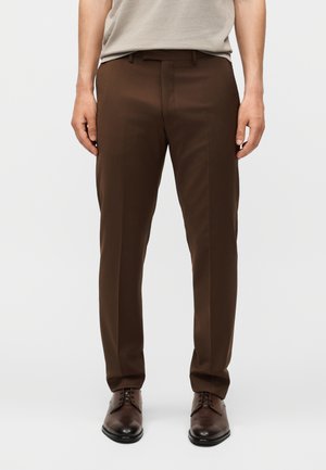 Mand iført mørkebrune habitbukser og matchende polerede brune læder habit sko, stående mod en ensartet lys baggrund.