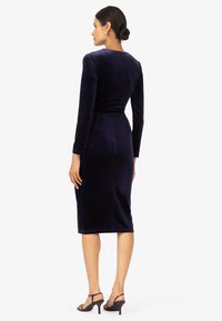 Navy blauwe fluwelen midi-jurk met lange mouwen, aansluitende silhoeette en getextureerde afwerking. Gestyled met zwarte bandjeshakken en een strakke haardracht.