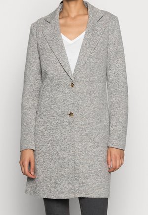 Vrouw draagt een lichtgrijs, getextureerd mantel van middellange lengte met twee gouden knopen over een wit T-shirt met V-hals en donkere broek.