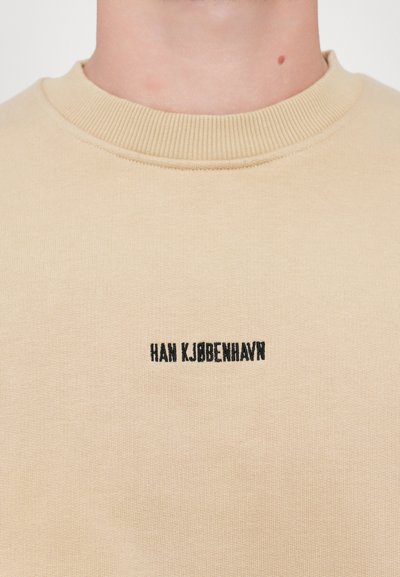 Beige bomulds-sweatshirt med ribbet crew-halsudskæring og sort broderet tekst, der viser "HAN KJØBENHAVN" på forsiden.