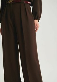 Pantalons en toile large marron, dotés de plis et d'une ceinture bordeaux brillante. Le design met en avant une coupe décontractée et un tombé élégant.